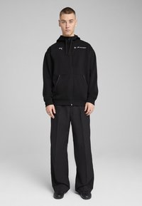 Hoodie noir zippé avec des accents violets, poches avant et détails de logo, associé à un pantalon noir ample. Design simple et dépouillé.