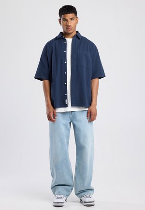 Navyblaues Kurzarm-Hemd mit Knöpfen und Tasche, kombiniert mit hellblauen, locker sitzenden Jeans und weißen Sneakern mit grauen Akzenten.