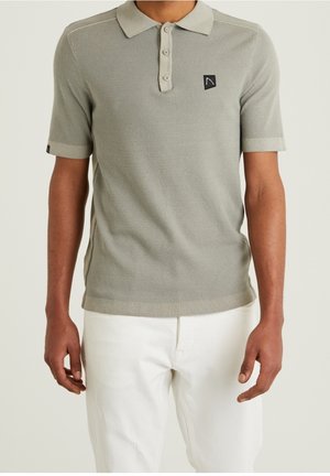 Jasnoszara teksturowana polo z krótkim rękawem, z kołnierzykiem i trzema guzikami, noszona z białymi spodniami przez stojącą osobę.