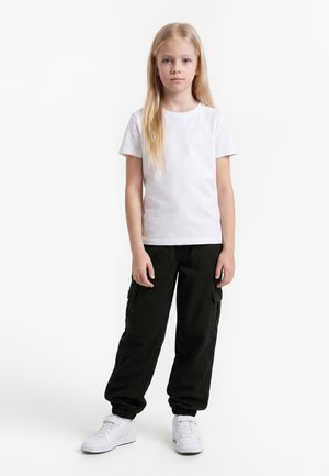 Fată tânără cu păr blond lung, purtând un tricou alb, pantaloni cargo negri și teniși albi, stând în fața unui fundal alb simplu.