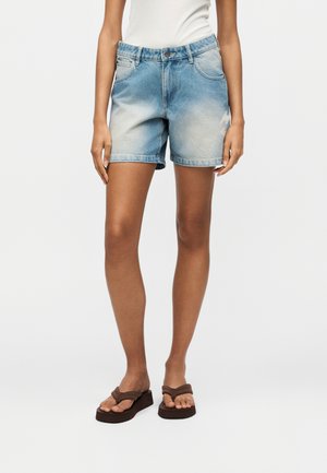 Γυναίκα που φοράει ανοιχτό μπλε τζιν σορτς με ξεθωριασμένο ξεβάμμα, λευκό τοπ και καφέ πλατφόρμες σαγιονάρες, στέκεται μπροστά σε λευκό φόντο.