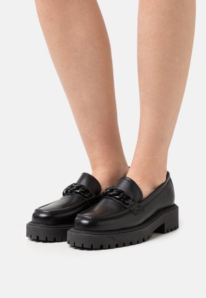 Mocassins en cuir noir avec une semelle noire épaisse, présentant un accent en chaîne sur le devant. Texture lisse avec un design de bout arrondi.