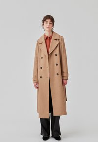 Cappotto lungo in misto lana marrone con bottoni doppi, ampi rever e tasche laterali, indossato sopra una camicia arancione e pantaloni neri.