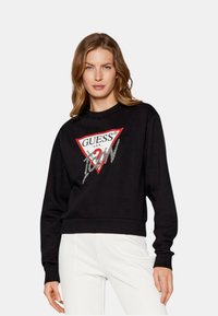 Sudadera negra con un logo triangular rojo que presenta el texto "GUESS" y acentos gráficos, de mangas largas y diseño recortado.
