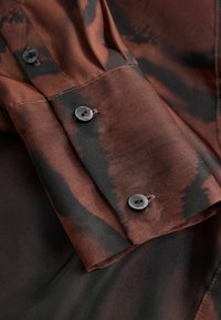 Ilga rankovė marškiniai, turintys turtingą rudos ir juodos spalvos raštą, su apvaliais juodais mygtukais ant rankovių, pagaminti iš lygaus audinio su blizgiu paviršiumi.