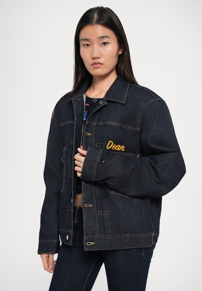 Veste en denim bleu foncé avec le prénom "Dean" brodé en jaune, dotée d'une fermeture à boutons, de poches et de détails de couture contrastants.
