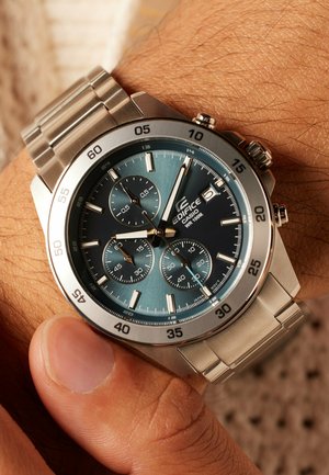 Silberne Edelstahluhr mit blauem Farbverlauf-Zifferblatt, drei Hilfszifferblättern und Leuchtmarkierungen. Stoppuhrfunktion und strukturierte Lünette-Details.