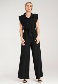 Czarny kombinezon z krótkimi, szerokimi rękawami, zapięciem na guziki z przodu, dwiema kieszeniami na piersi i paskiem do wiązania w talii. Luźny, szeroki fason nogawek.