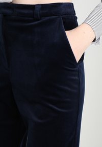 Námořnické modré sametové kalhoty s hladkou strukturou, vybavené bočními kapsami a přiléhavým pasem. Žádné viditelné kovové komponenty nebo vzory.