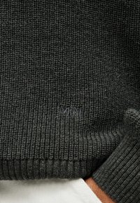 Μανίκι και τελείωμα σκούρου γκρι ριμπ πλεκτού πουλόβερ πάνω από λευκό ύφασμα, με διακριτική ανάγλυφη κεντητική κοντά στο τελείωμα.