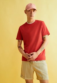 Rode katoenen T-shirt met korte mouwen, ronde hals, gedragen met lichtbeige shorts. Roze pet met logo. Achtergrond is een gele muur.