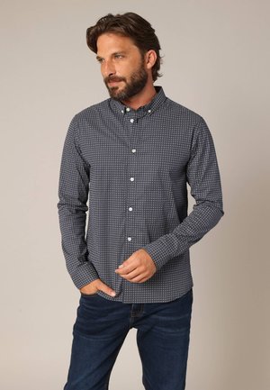 Een knoopsluiting shirt in marineblauwe stof met een klein wit geometrisch patroon. Heeft een klassieke kraag en lange mouwen, gecombineerd met blauwe jeans.