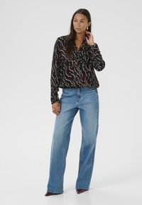 Zwarte blouse met een wit en rood abstract patroon, gecombineerd met lichtblauwe wijde jeans. Het model draagt puntige rode hakken en heeft lang haar.