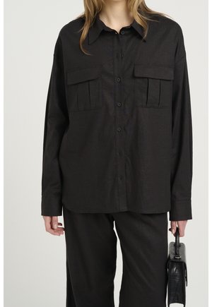 Kobieta ubrana w luźną czarną koszulę na guziki z dwiema kieszeniami na piersi oraz pasujące czarne spodnie, trzymająca małą czarną torebkę.