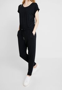 Frau in einem schwarzen kurzärmligen Overall mit Kordelzug und Taschen, kombiniert mit weißen Sneakers, vor einem schlichten Hintergrund stehend.