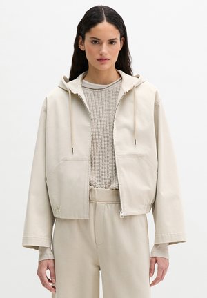 Kobieta z długimi ciemnymi włosami, ubrana w beżową kurtkę na zamek z kapturem, nałożoną na teksturowany dzianinowy sweter oraz pasujące beżowe spodnie, stojąca na białym tle.