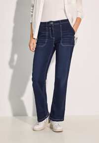 Jeans larghi di colore blu scuro con cuciture a contrasto, due tasche anteriori e zip, abbinati a sneaker bianche e una camicia chiara.