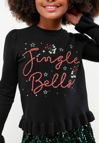 Melnas džemperis ar "Jingle Bells" sarkanos diegos un dekoratīvām pērlītēm. Ar pūkainām piedurknēm un viļņotu apakšmalu. Zem tā zaļa kleita ar pērlītēm.