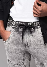 Manos en los bolsillos de pantalones de chándal gris con efecto ácido y cordón negro, combinados con una camiseta blanca y una chaqueta con cremallera negra.