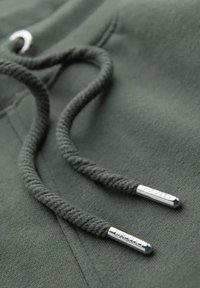 Olivgrüner Kapuzenpulloverstoff mit strukturierter Oberfläche; dicke, geflochtene Kordeln mit silbernen Metallenden, die eingravierte Buchstaben aufweisen.