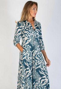 Długa sukienka koszulowa w kolorze teal z białymi abstrakcyjnymi wzorami. Posiada kołnierzyk, półdługie rękawy oraz boczne kieszenie, wykonana z miękkiego materiału.