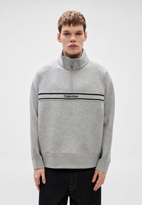 Γκρι φούτερ με φερμουάρ και ψηλό γιακά, με οριζόντια μαύρη λωρίδα και το λογότυπο "Calvin Klein" στο στήθος. Μαλακή υφή.