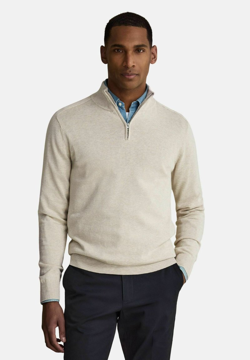 Homme portant un pull beige à fermeture éclair au quart sur une chemise bleue à boutons et un pantalon sombre, debout avec une main dans la poche.