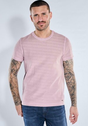 Mann mit kurzen Haaren und tätowierten Armen, der ein hellrosa kurzärmeliges Hemd und blaue Jeans trägt, steht vor einem schlichten hellen Hintergrund.