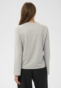 Sieviete ar garām brūnām matiem, tērpta gaiši pelēkā garajā piedurknē kreklā un melnās biksēs, redzama no mugurpuses uz vienkārša fona.