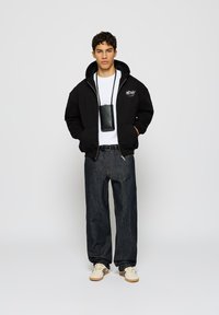 Svart hettegenser med grafisk logo, hvit t-skjorte, mørke jeans og kremfargede sneakers. Liten svart telefonveske båret rundt halsen.