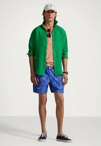 Πράσινο λινό πουκάμισο με κουμπιά πάνω από μια ρίγα πορτοκαλί και λευκό T-shirt, μπλε λουλουδάτα σορτς και ναυτικά παπούτσια. Τα αξεσουάρ περιλαμβάνουν ένα λευκό καπέλο.