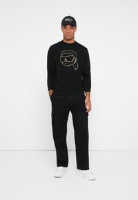 Uomo in piedi contro uno sfondo bianco, indossa un felpa nera con una faccia grafica, pantaloni neri, berretto nero e sneakers bianche. Mani nelle tasche.