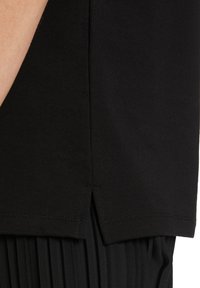 Camisa negra de manga corta con una abertura lateral, hecha de tela suave. El dobladillo presenta una costura sencilla, acompañado de tela negra plisada debajo.