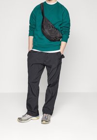 Zelená mikina, černé pracovní kalhoty a šedé tenisky. Černá taška přes rameno je pohodlně položena. Materiál vypadá hladký a lehce lesklý.