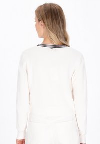 Vrouw met blond haar, gekleed in een witte trui met een zwart en rode gestreepte kraag, gezien van achteren tegen een witte achtergrond.