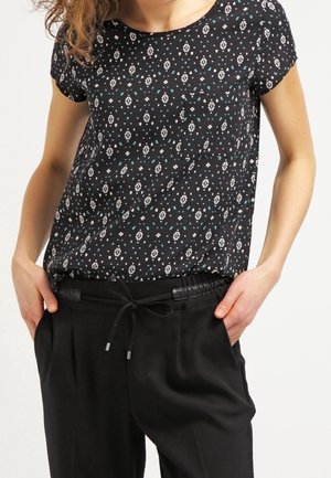 Kvinde iført en sort kortærmet mønstret bluse ind tucked i sorte bukser med snøre, hænderne hviler i lommerne mod en ensfarvet baggrund.