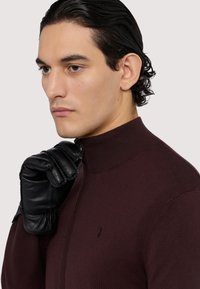 Bordový pletený svetr s vysokým límcem, zdobený malým logem. Modelka má na sobě černé kožené rukavice, které zvýrazňují texturu a kontrast.