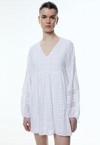 Weißes, strukturiertes Kleid mit V-Ausschnitt, langen Ärmeln und Spitzenverzierungen an den Bündchen. Lockere Passform, geraffte Taille, karriertes Muster überall.