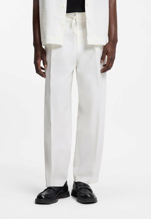 Mand iført løse hvide teksturerede bukser med snøre i taljen og sorte læder loafers, stående mod en hvid baggrund.