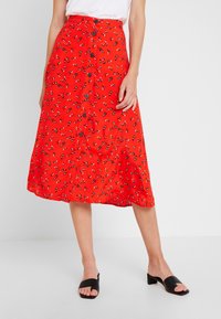 Jupe midi rouge avec des motifs floraux noirs et blancs dispersés, dotée de boutons sur le devant et d'un tissu fluide. Associée à des sandales à glissière noires.
