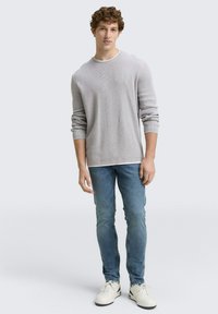 Hellgrauer gerippter Pullover mit langen Ärmeln und Rundhalsausschnitt, kombiniert mit engen blauen Jeans und weißen Sneakers. Einfaches, lässiges Design.