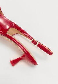 Scarpa con tacco alto in vernice rossa, con design slingback, caratterizzata da un tacco sottile e un piccolo dettaglio in oro sulla cinghia.