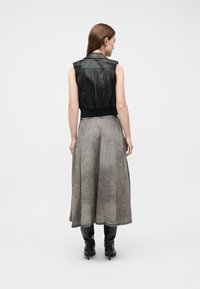 Γυναίκα που φοράει αμάνικο μαύρο δερμάτινο γιλέκο, γκρι μίντι φούστα σε γραμμή Α και μαύρες μπότες με τακούνι, στέκεται πλάτη σε λευκό φόντο.