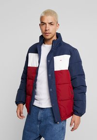 Veste rembourrée avec des blocs de couleurs bleu marine, blanc et rouge. Texture douce, boutons-pression, col haut. Comprend un logo sur la poitrine gauche et des poignets élastiques.