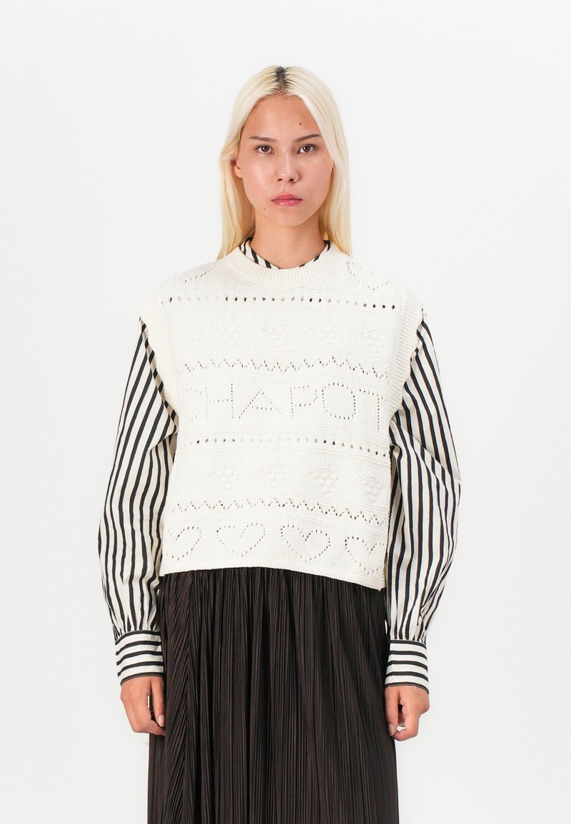 White knitted crop sweater with textured patterns and openwork. Layered over a black pleated skirt with vertical white stripes.