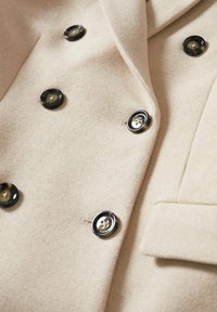 Manteau beige croisé avec des boutons à quatre trous bordés de noir et une poche à rabat frontale visible sur le côté droit.