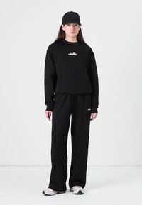 Svart treningsdrakt bestående av en cropped hettegenser og vide bukser. Begge plagg viser merkevaren sin logo i hvitt, kombinert med en matchende svart caps.
