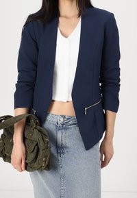 Navy blejzr s úzkým střihem, cropped siluetou a kapsami na zip. V kombinaci s bílým žebrovaným topem a sukní z denimu s vysokým pasem. Olivově zelená taška.