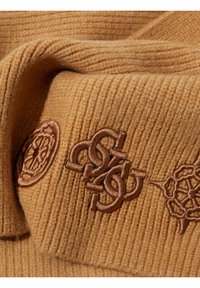 Tessuto a maglia a coste marrone con motivi ricamati, inclusi disegni circolari e intrecciati, che mostrano una texture morbida e un colore caldo.