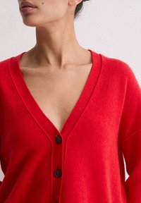 Czerwony kardigan z dekoltem w serek, wykonany z miękkiej tkaniny, z trzema czarnymi guzikami z przodu i luźnym krojem.